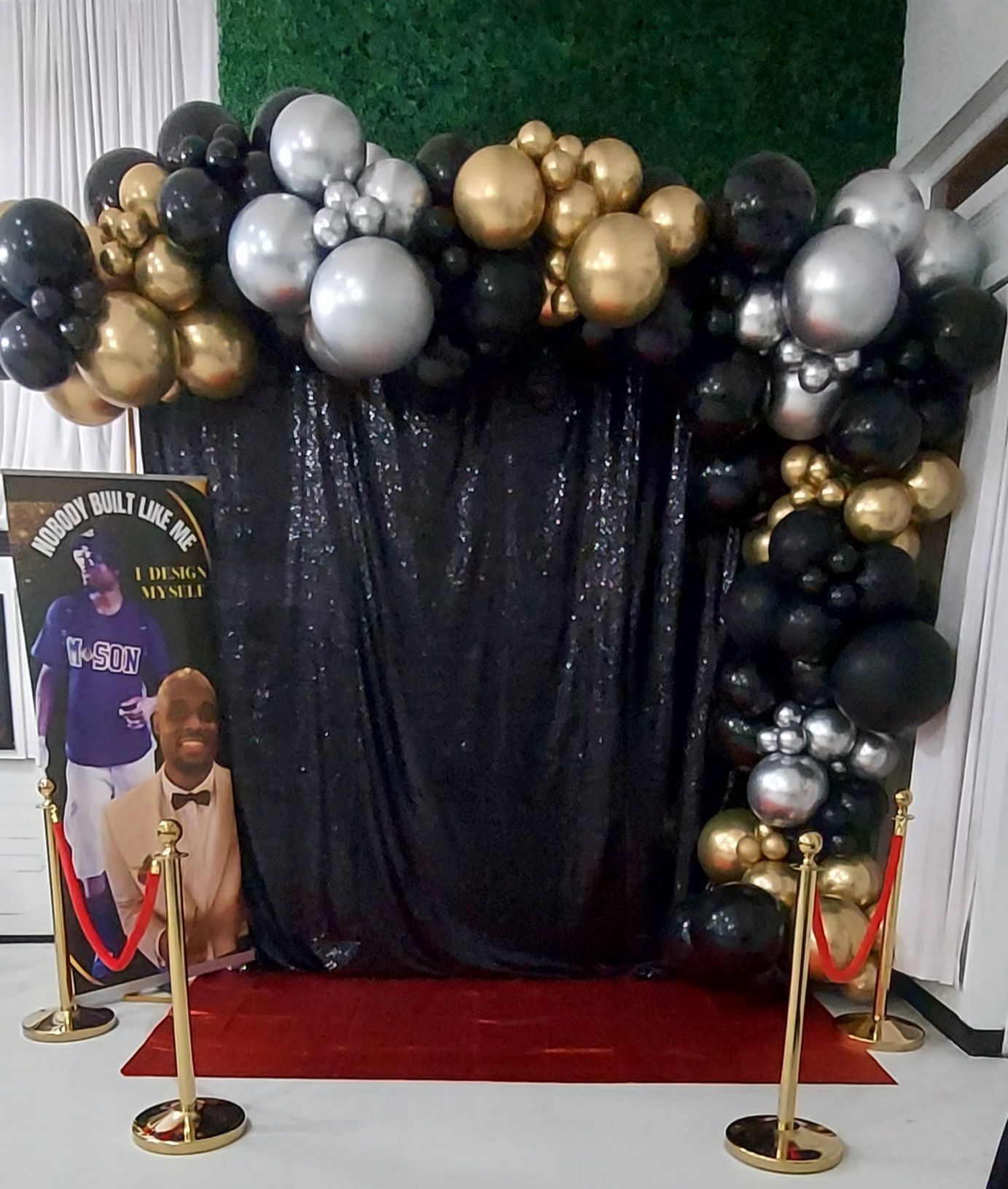 A black and gold balloon arch frames a sequined backdrop with a red carpet and gold stanchions at a formal event.