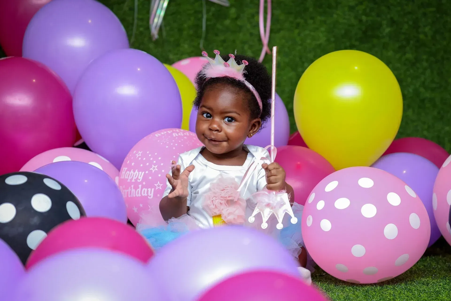 A smiling Black toddler wearing a crown, surrounded by colorful balloons.