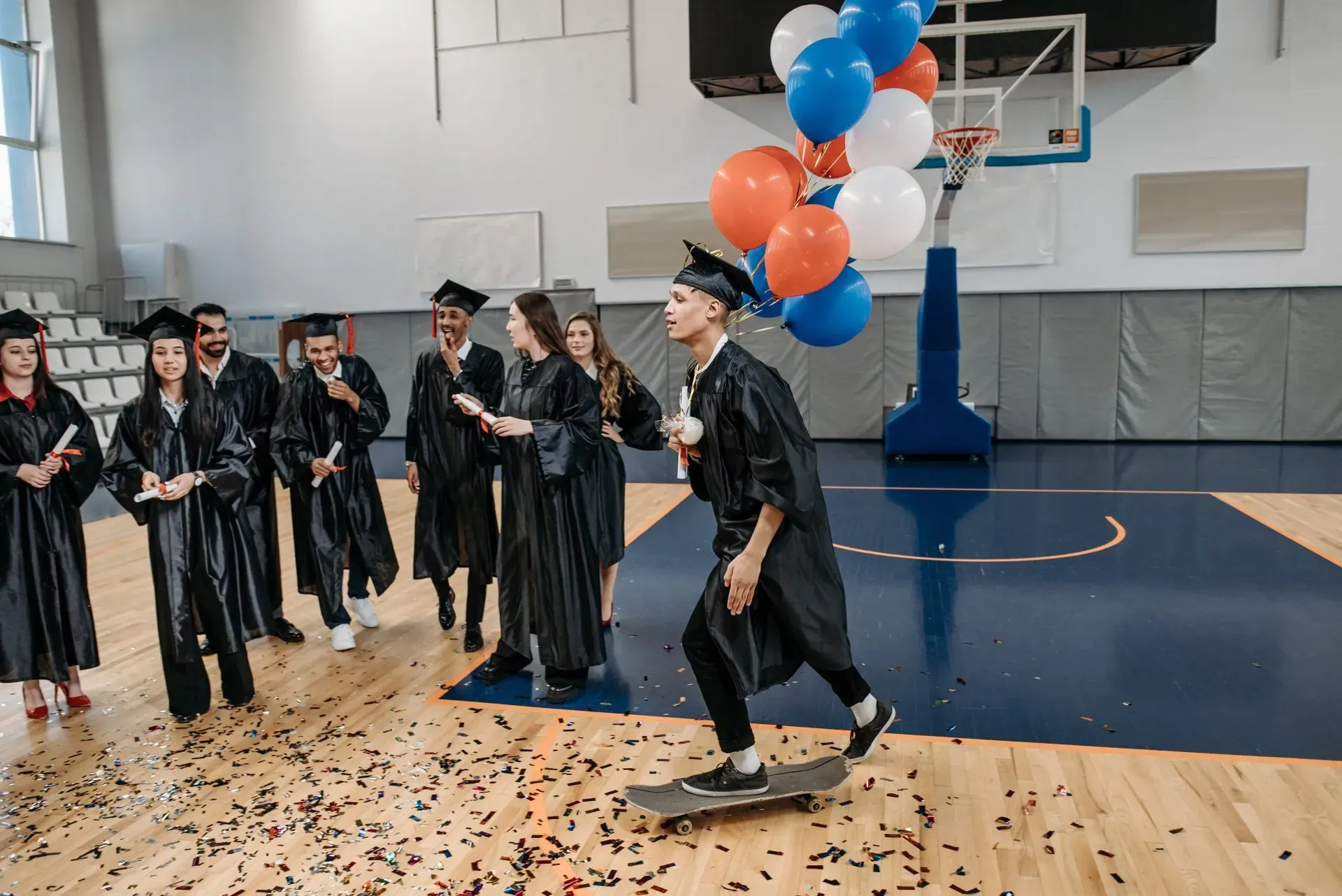 A graduate on a skateboard holding balloons, cheered on by classmates in a gym.