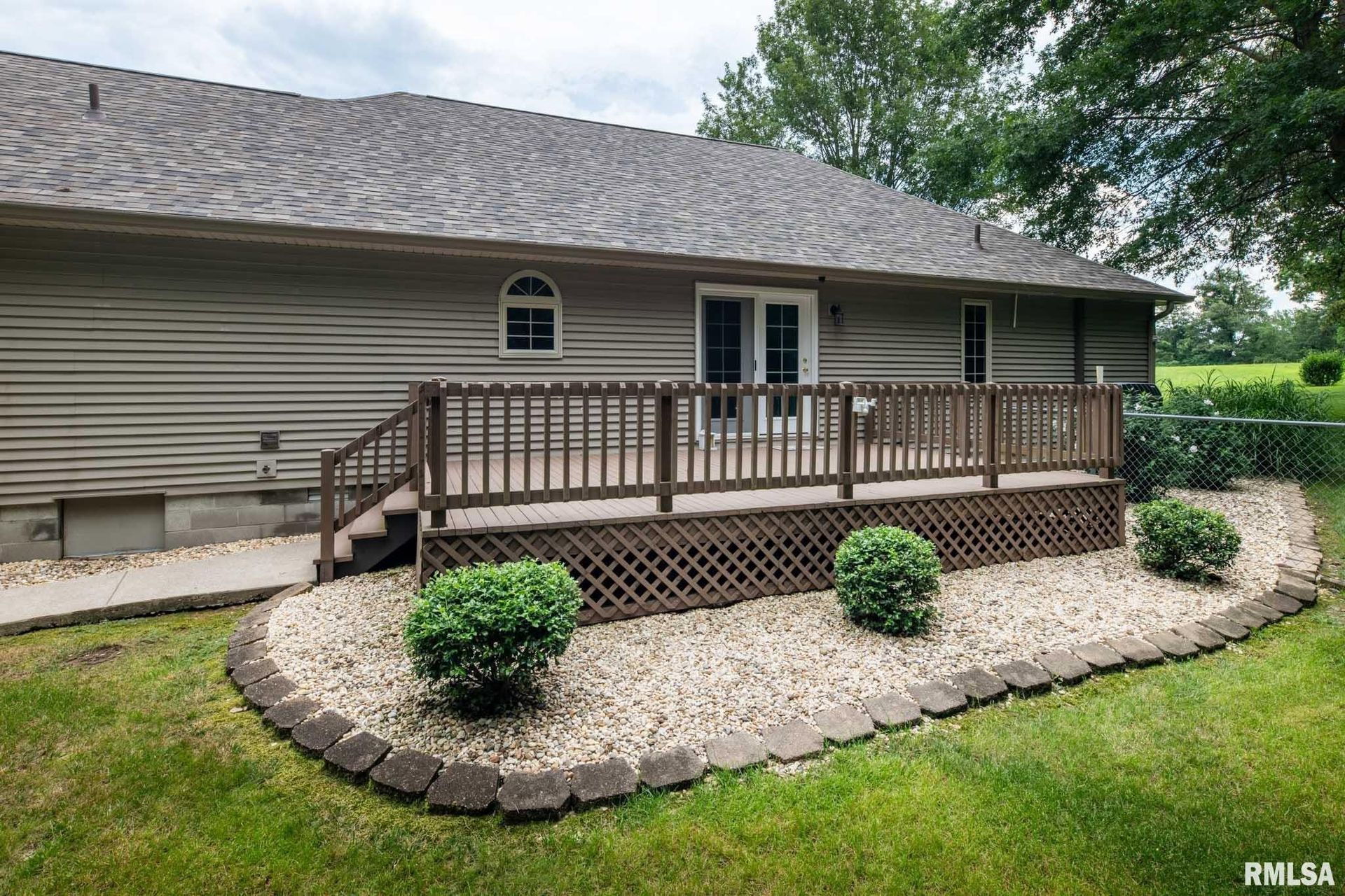 House with raised wooden deck and stairs, set in a landscaped yard with stone edging and shrubs