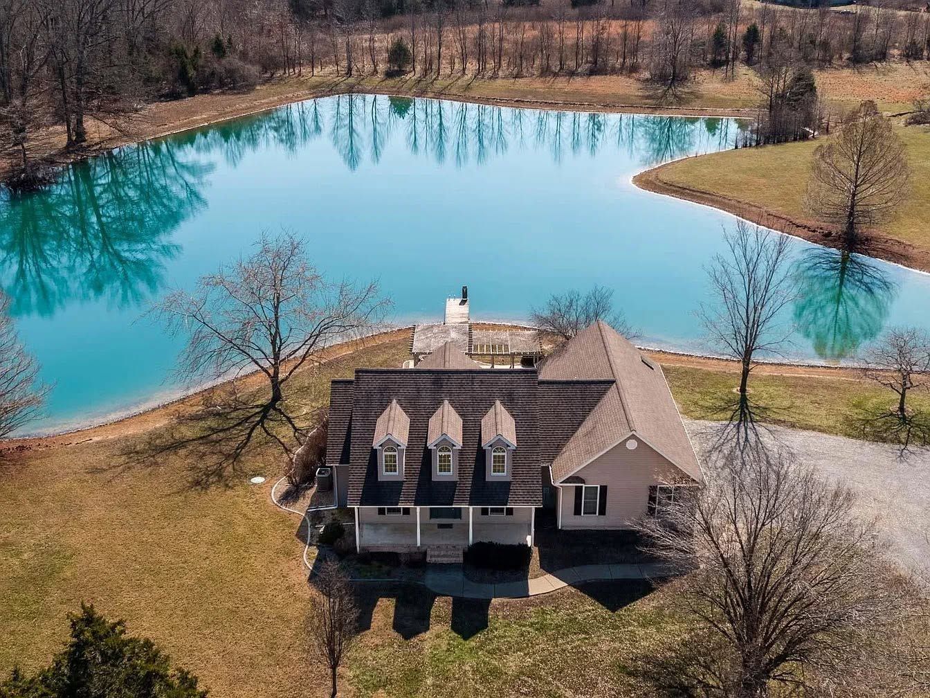 Aerial view of a house beside a bright turquoise lake surrounded by bare trees.