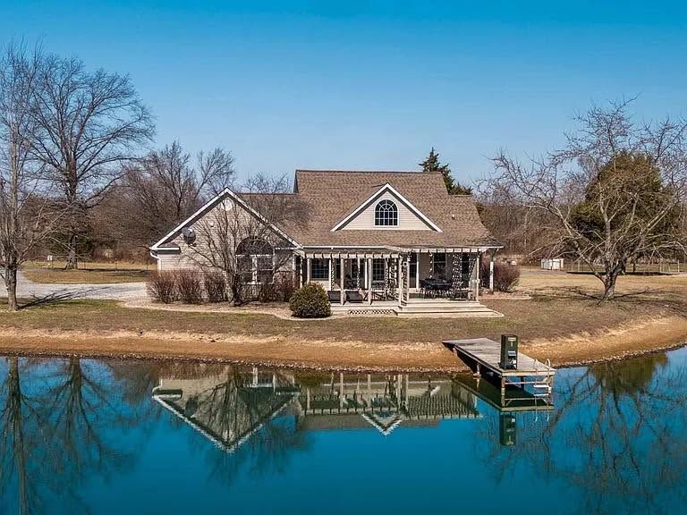 Lakefront house with tan roof and porch, reflected in calm water on a clear day