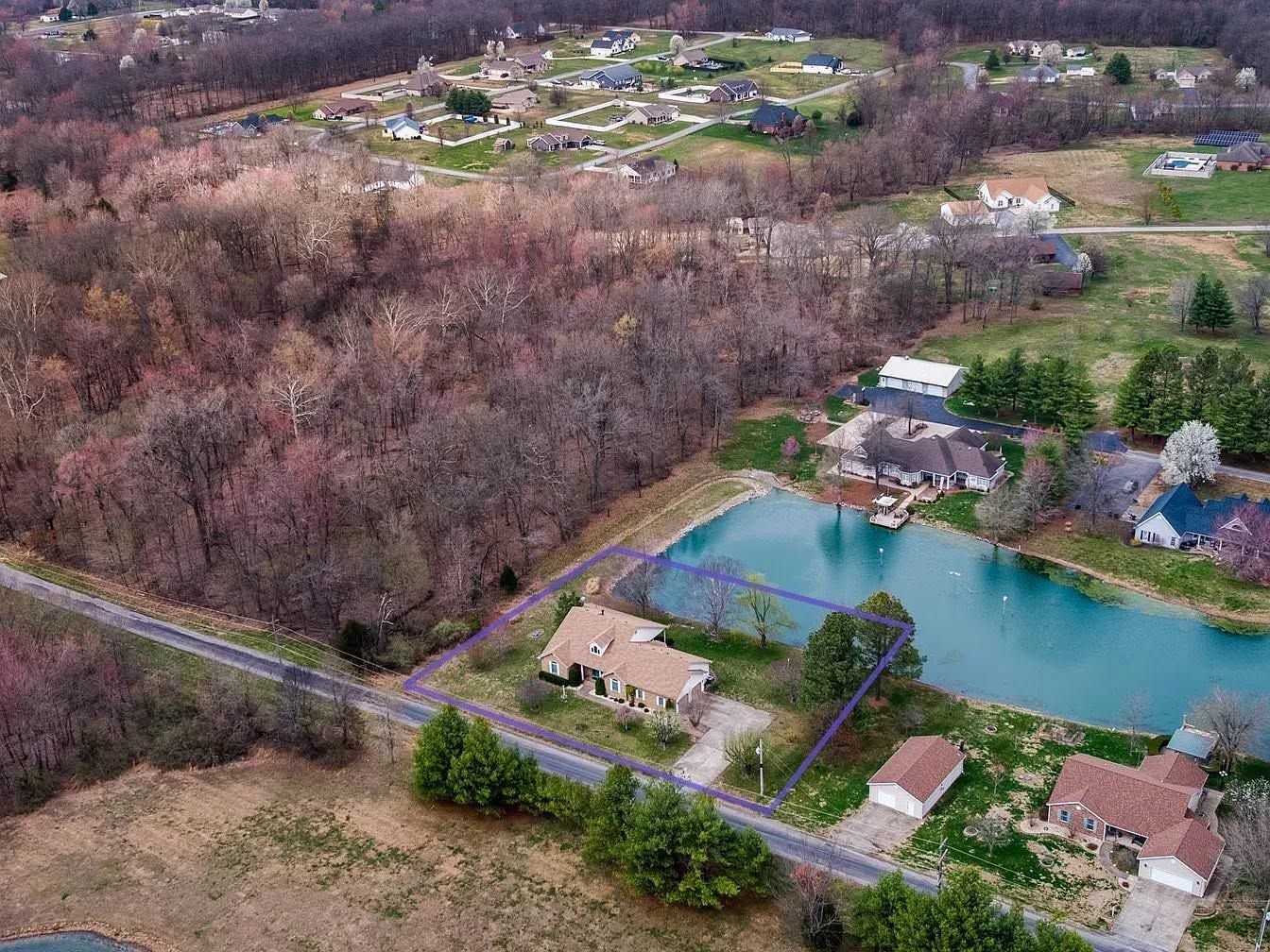 Aerial view of suburban homes beside a blue pond and wooded area, with roads and fields nearby