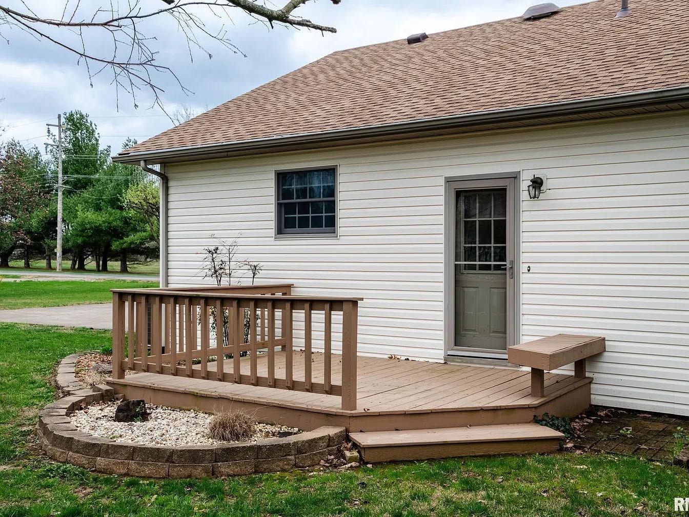 Small white house exterior with a wooden deck, railing, and side door in a grassy yard.