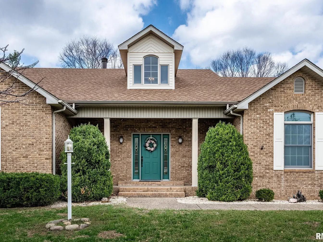 Brick house with teal front door, dormer window, and neatly trimmed shrubs under a clear sky