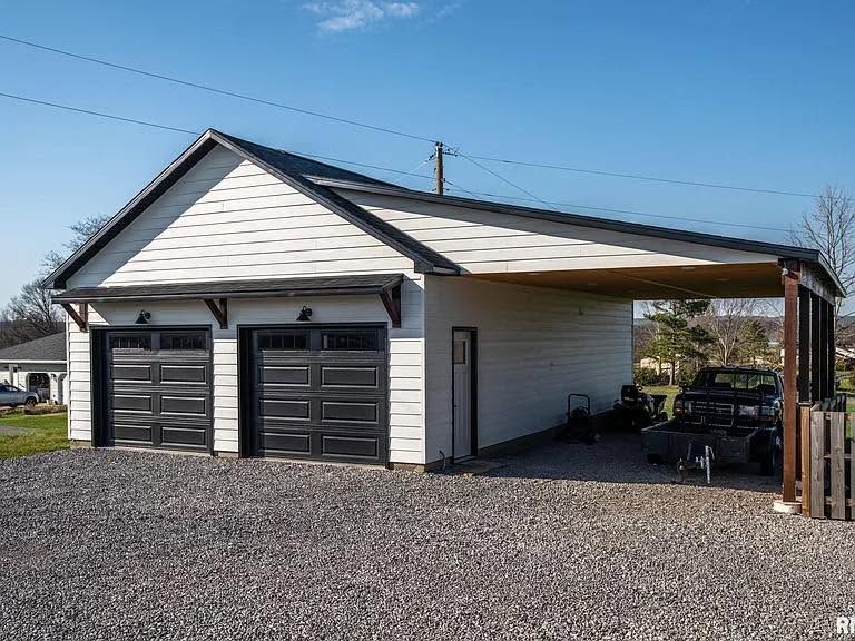 White detached garage with two black doors and a carport on the right, viewed from a gravel driveway