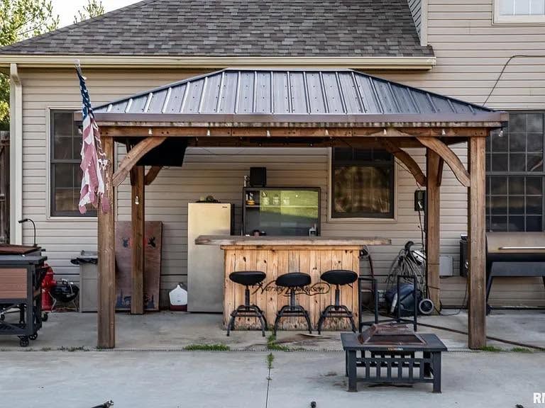 Covered backyard patio with wooden bar, stools, grill, and an American flag beside a house