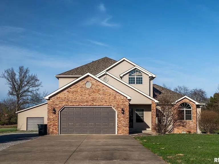 Suburban two-story brick house with attached garage, driveway, and green lawn under a clear blue sky