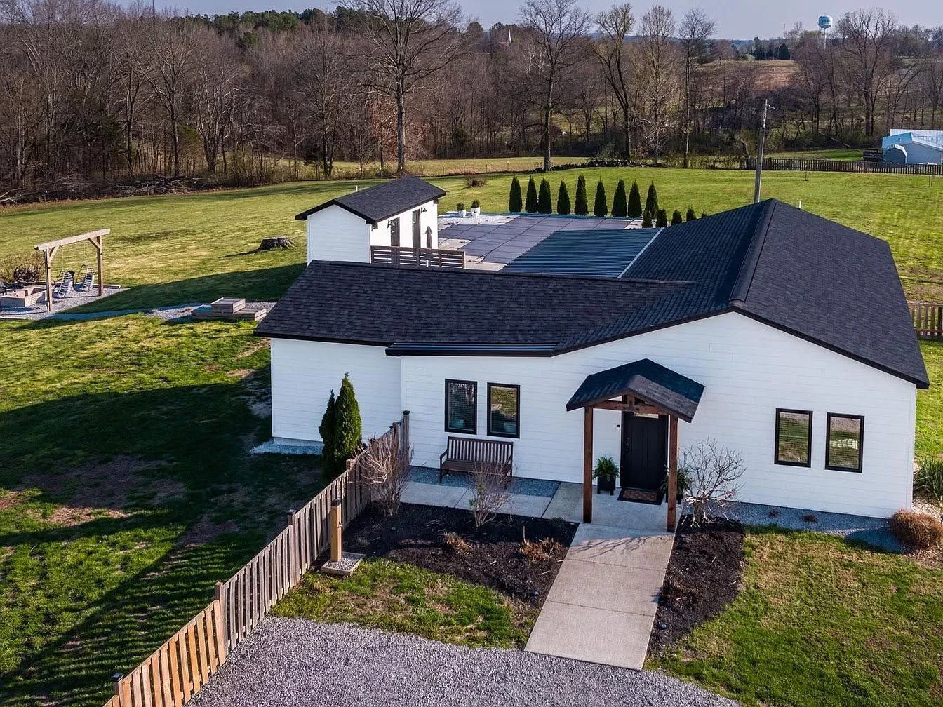 White house with dark roof and fenced yard in a grassy rural area, aerial view