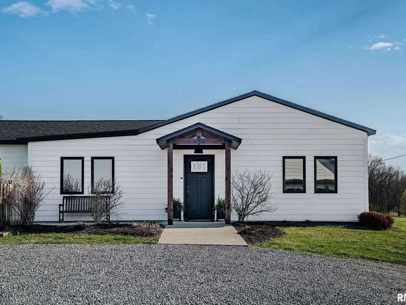 Small white house with black trim, front door, windows, and gravel yard under a blue sky