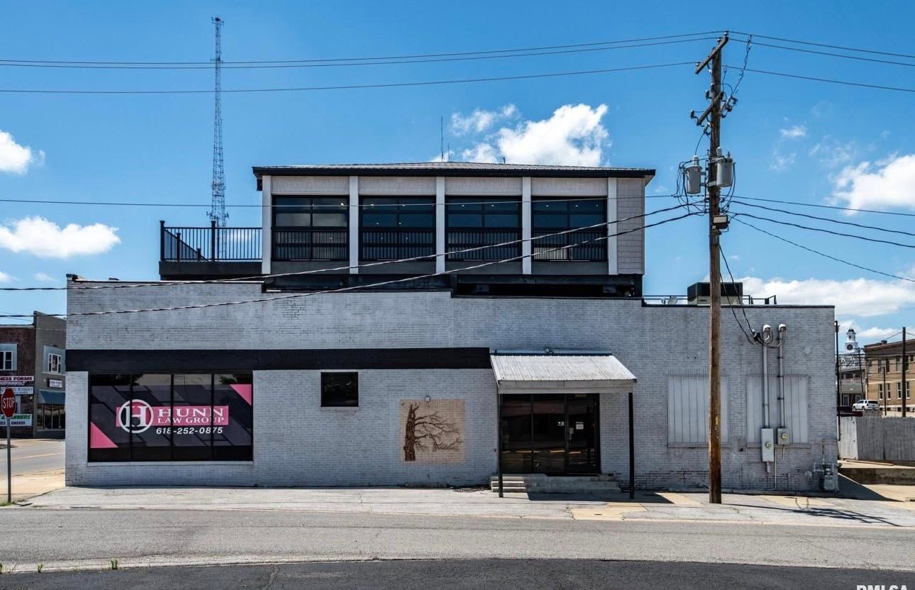 Small white commercial building with a dark storefront under a bright blue sky, seen from across the street