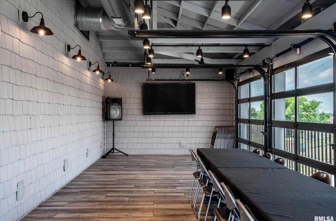 Outdoor covered patio with wood floor, white brick walls, TV, and long dining table with bar stools