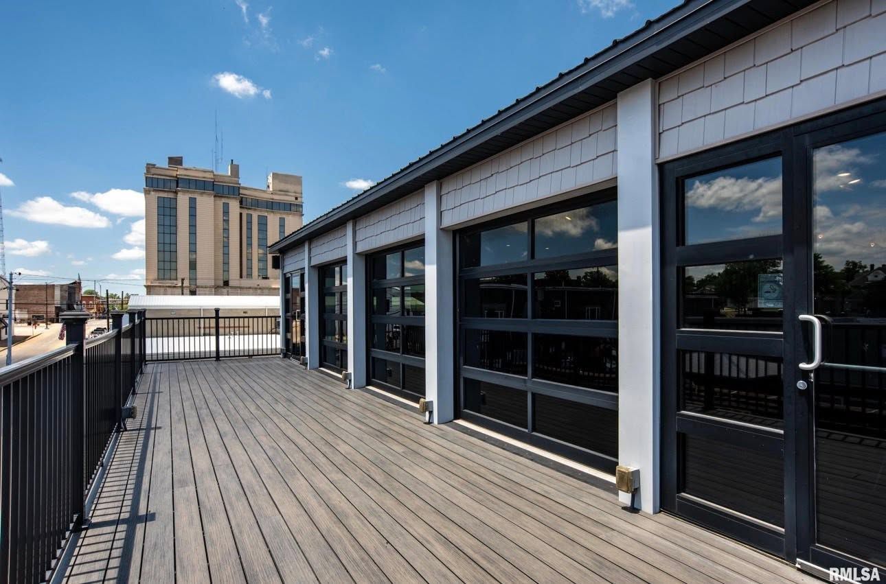 Rooftop wooden deck beside a building with large windows and city buildings in the distance.