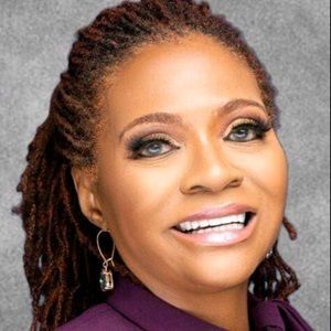 A woman with dreadlocks is smiling and wearing a purple shirt and earrings.