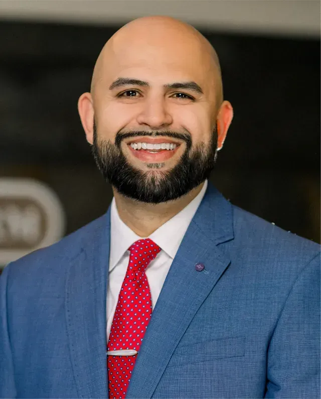A bald man with a beard is wearing a blue suit and red tie.
