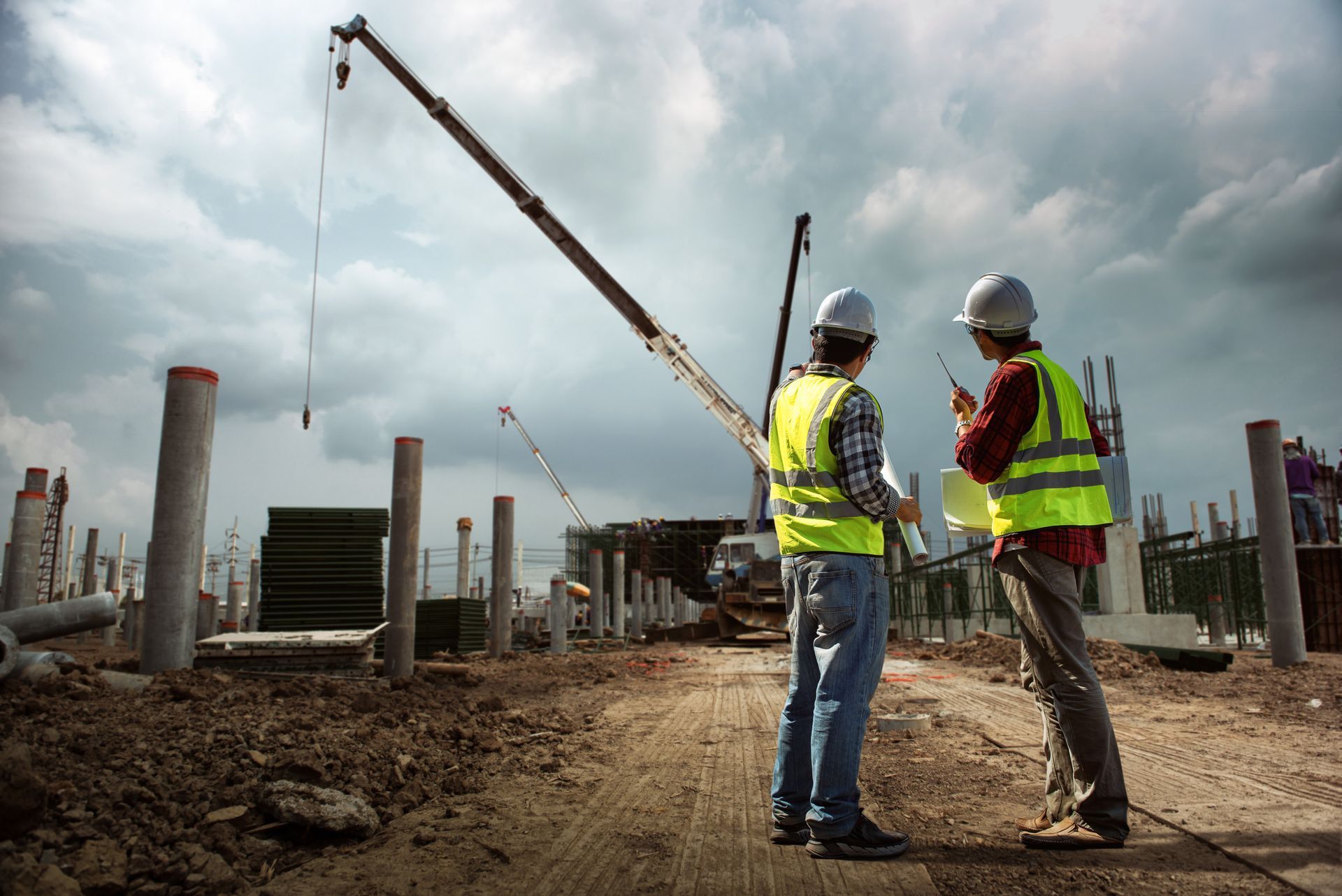 Two construction workers are standing on a construction site looking at a crane. | CA - United Nationwide Security Services Two construction workers are standing on a construction site looking at a crane. | CA - United Nationwide Security Services