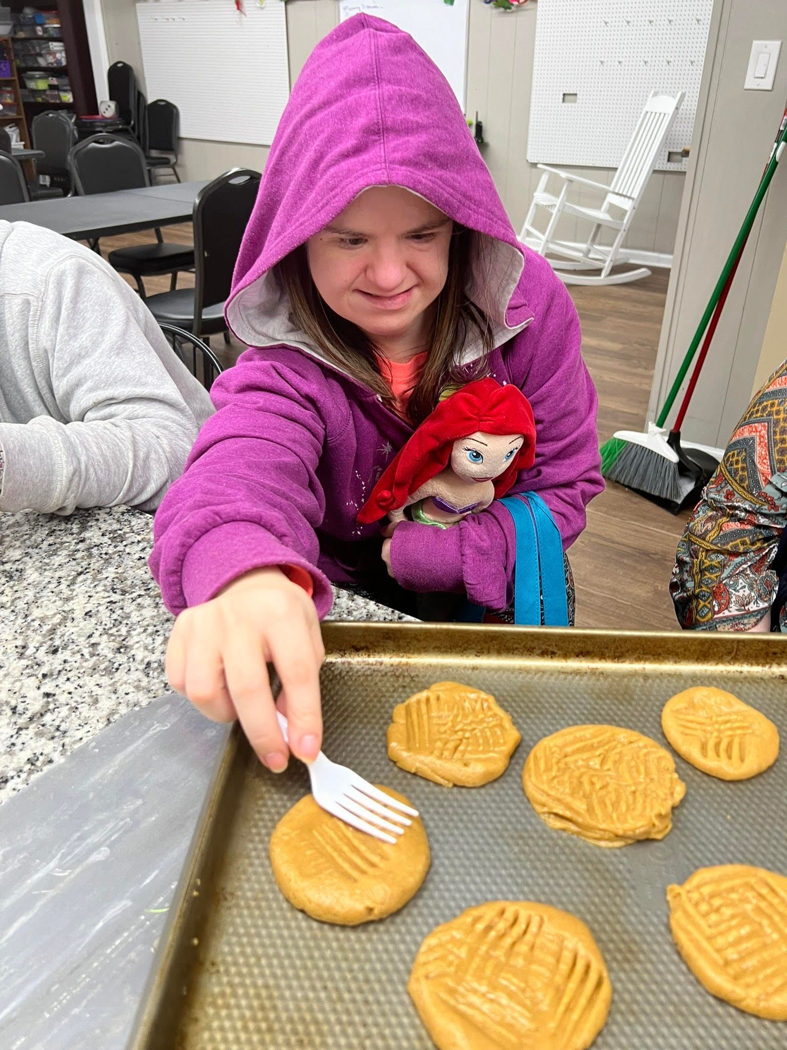 A girl in a purple jacket is holding a stuffed animal while cutting cookies with a fork.
