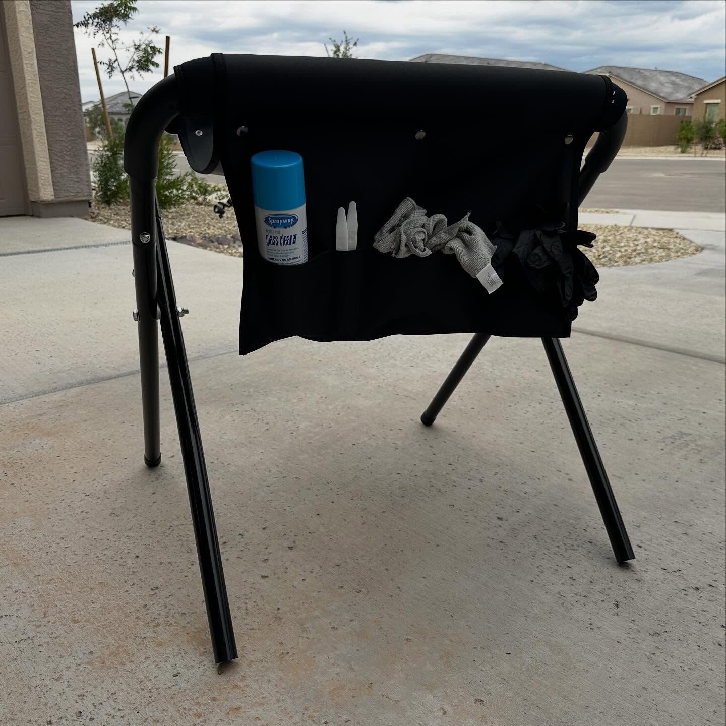 Black folding chair on a concrete driveway with a blue cup and gray gloves in the seat pocket.