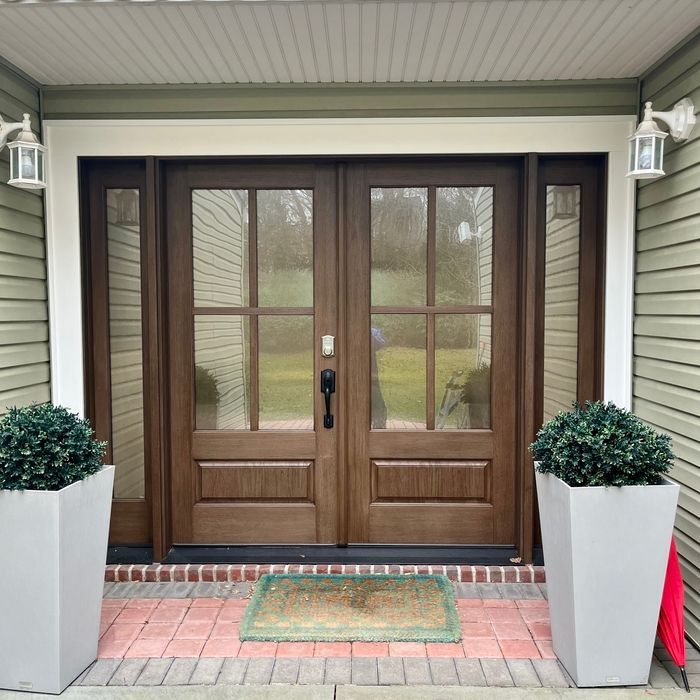 Brown double doors with glass panes, flanked by white planters with greenery and two wall lights.