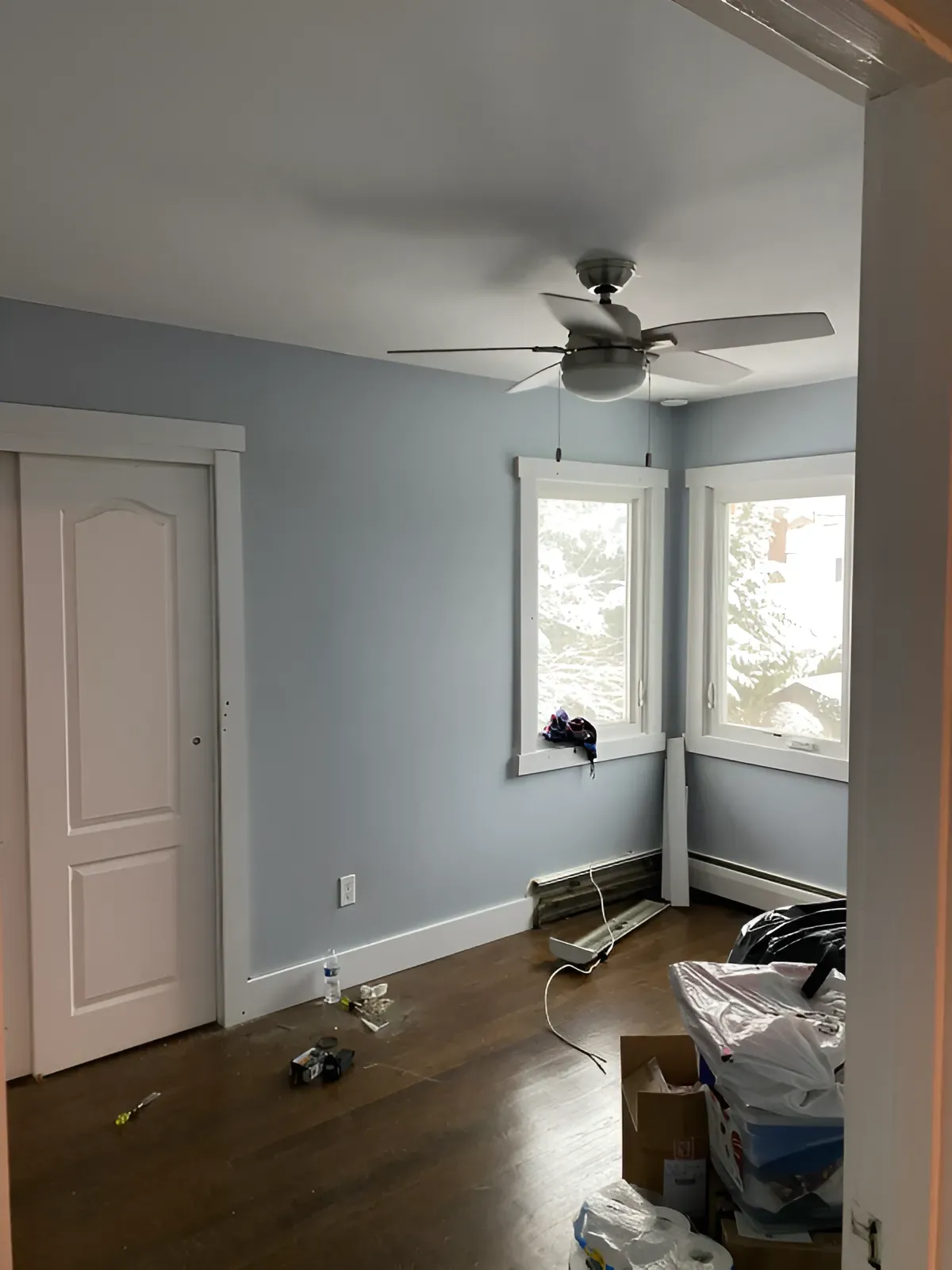 Empty room with blue walls, dark wood floor, and white trim. Ceiling fan. Door and windows visible.
