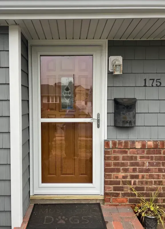 Exterior view of a front door. A white screen door covers a brown door. The house has gray siding and a brick base.