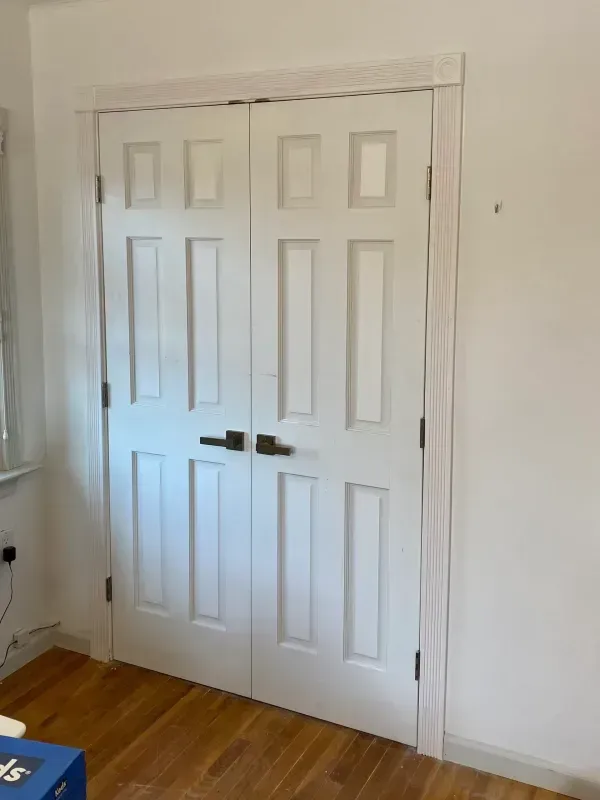 White double doors with dark handles and light trim, set in a light-walled room with wood flooring.