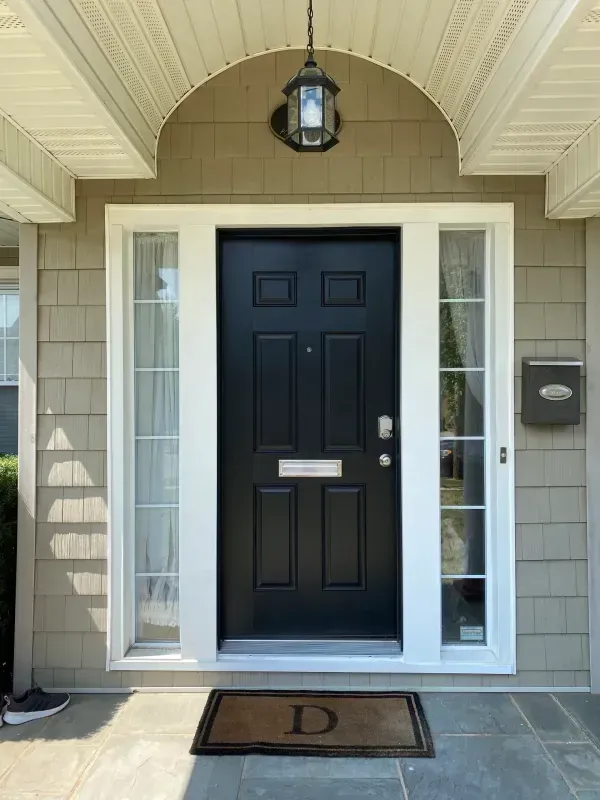 Black front door with sidelights, under a beige archway, features a welcome mat with the letter "D."