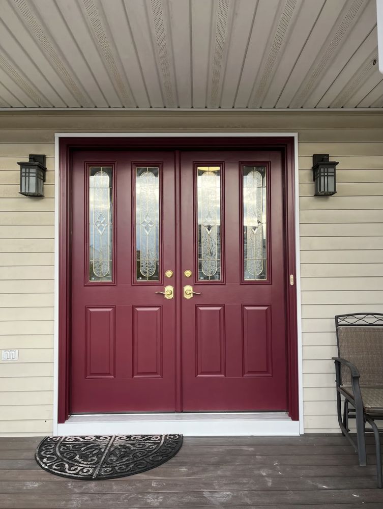 Double maroon front doors with glass panels and gold hardware; flanked by black sconces on a tan house.