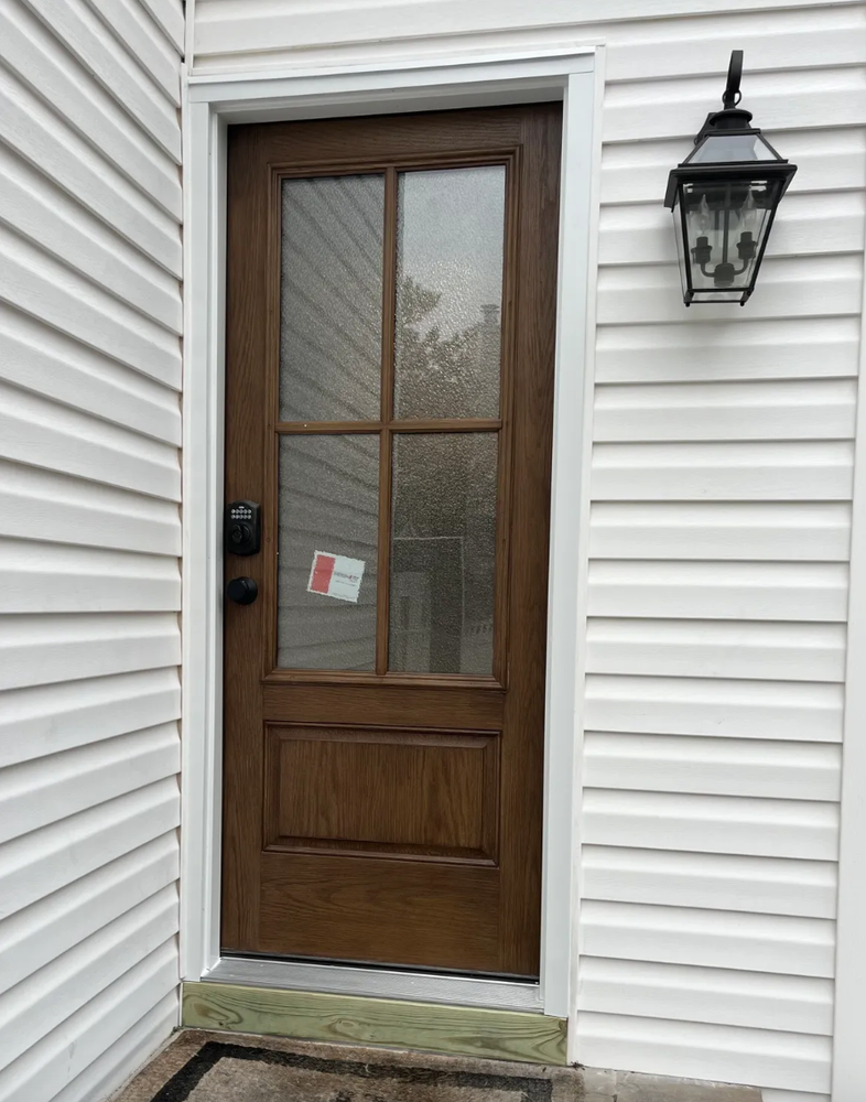Wooden door with glass panes and black hardware, surrounded by white siding, with a lantern on the right.