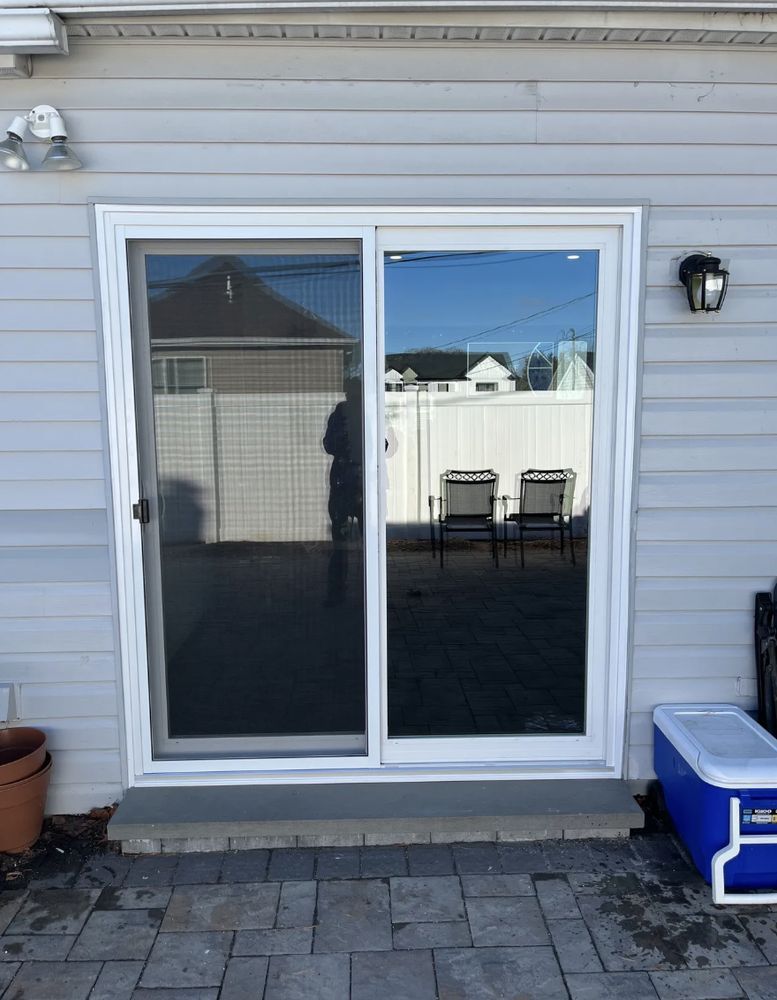 Sliding glass door, white frame, gray siding, patio, two chairs visible through window reflection.