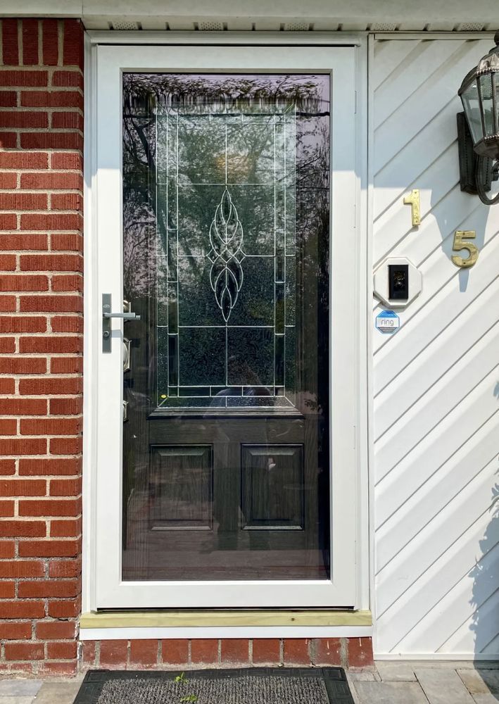 Front entrance with a white-framed glass storm door beside a brick wall and wall-mounted lantern.