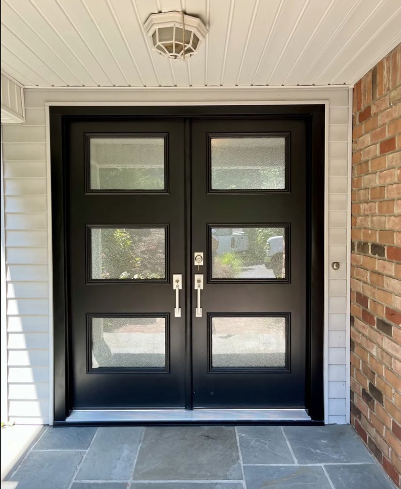 Black double front doors with rectangular windows, under a white ceiling fixture, on a stone patio.