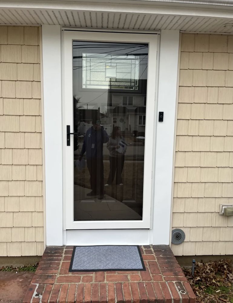 Glass front door with a white frame, reflective with two people visible, set in a brick entryway.