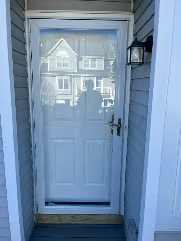 Front door with glass storm door reflecting a house across the street, white siding and porch light.