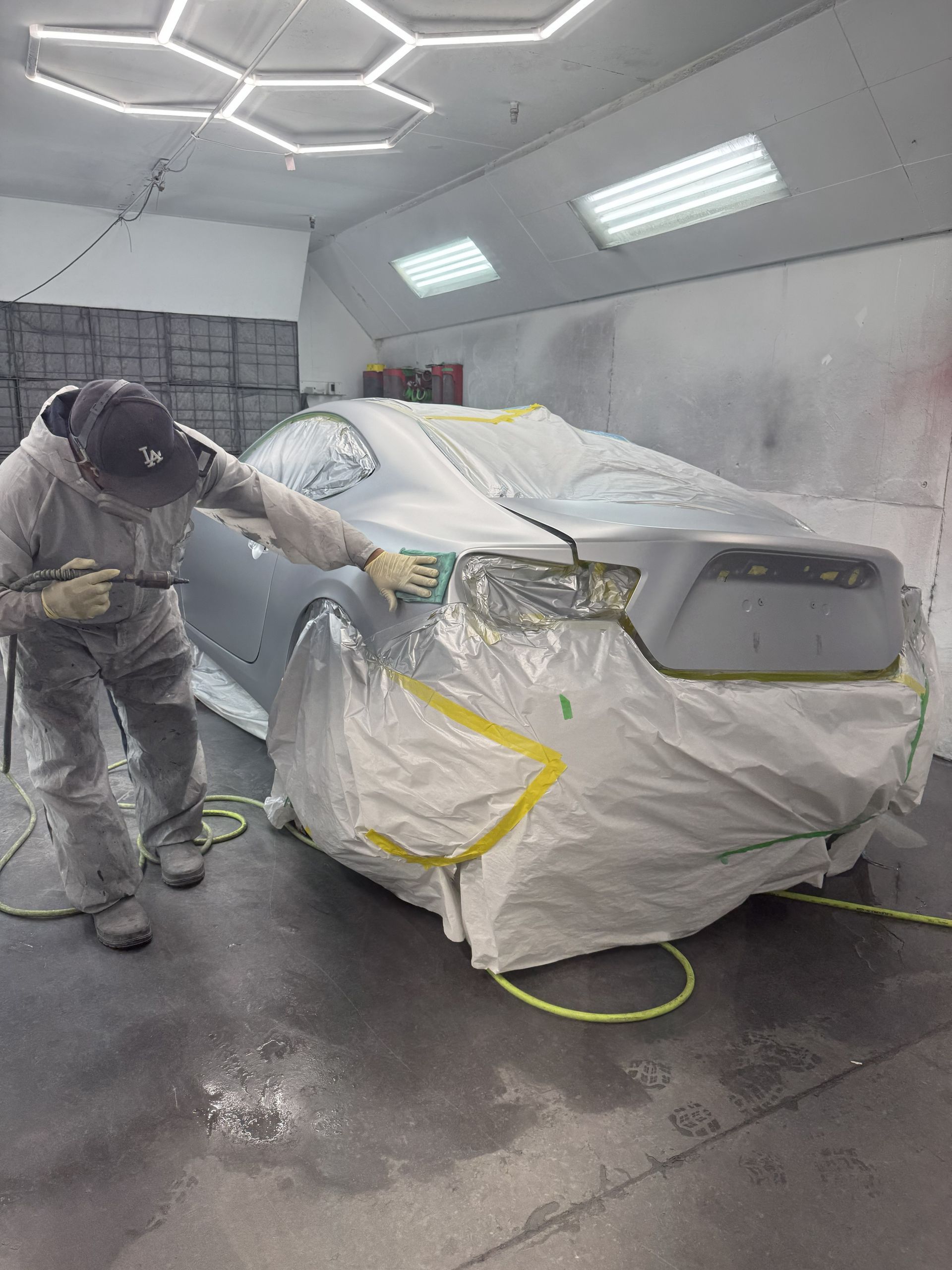 A person in a protective suit uses a spray gun to paint a grey car inside a well-lit automotive spray booth.