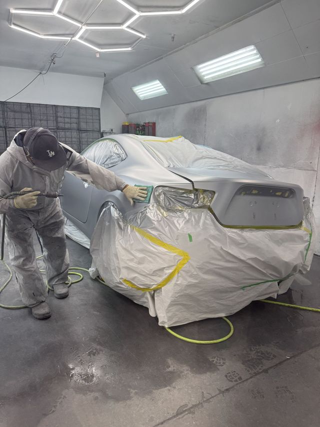 A person in a protective suit uses a spray gun to paint a grey car inside a well-lit automotive spray booth.