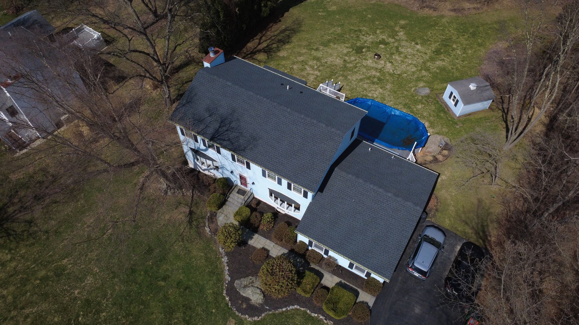Aerial view of a white house with a dark roof, driveway, parked car, and wooded yard.