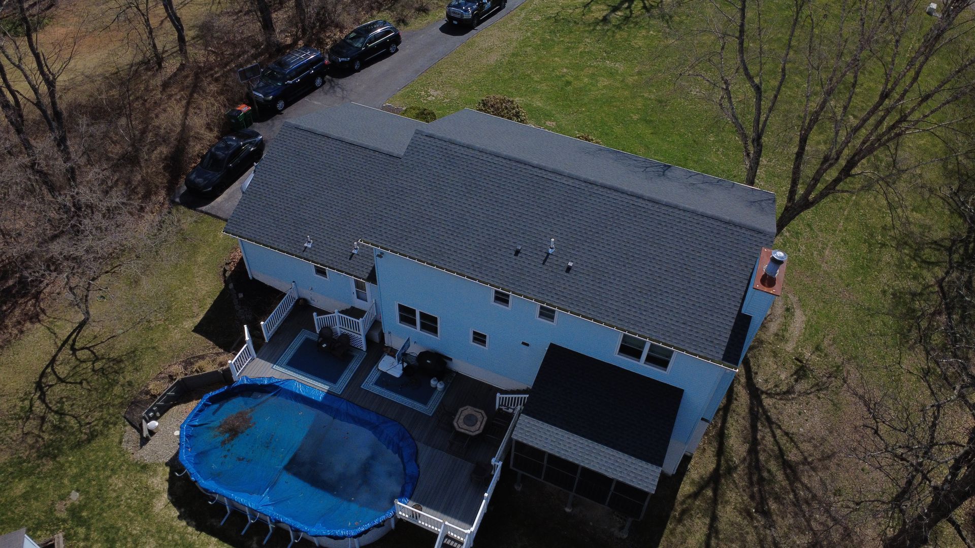 Aerial view of a house with a dark roof, backyard deck, and blue oval pool surrounded by trees.
