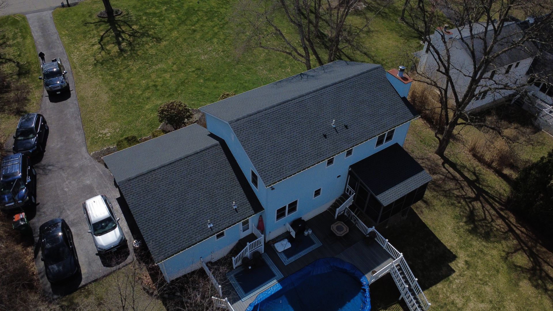 Aerial view of a blue house with dark roofs, driveway cars, and a backyard pool.
