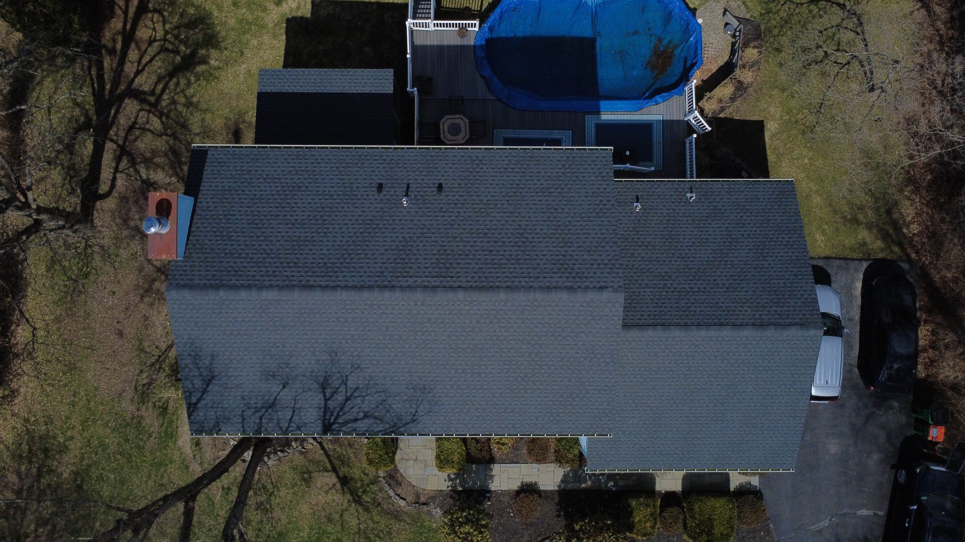 Aerial view of a gray-roofed house with a driveway, trees, and a blue above-ground pool.
