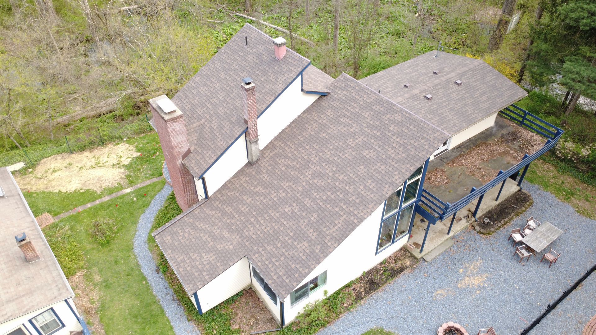 Aerial view of a white house with brown roof, blue deck, and gravel driveway surrounded by trees.