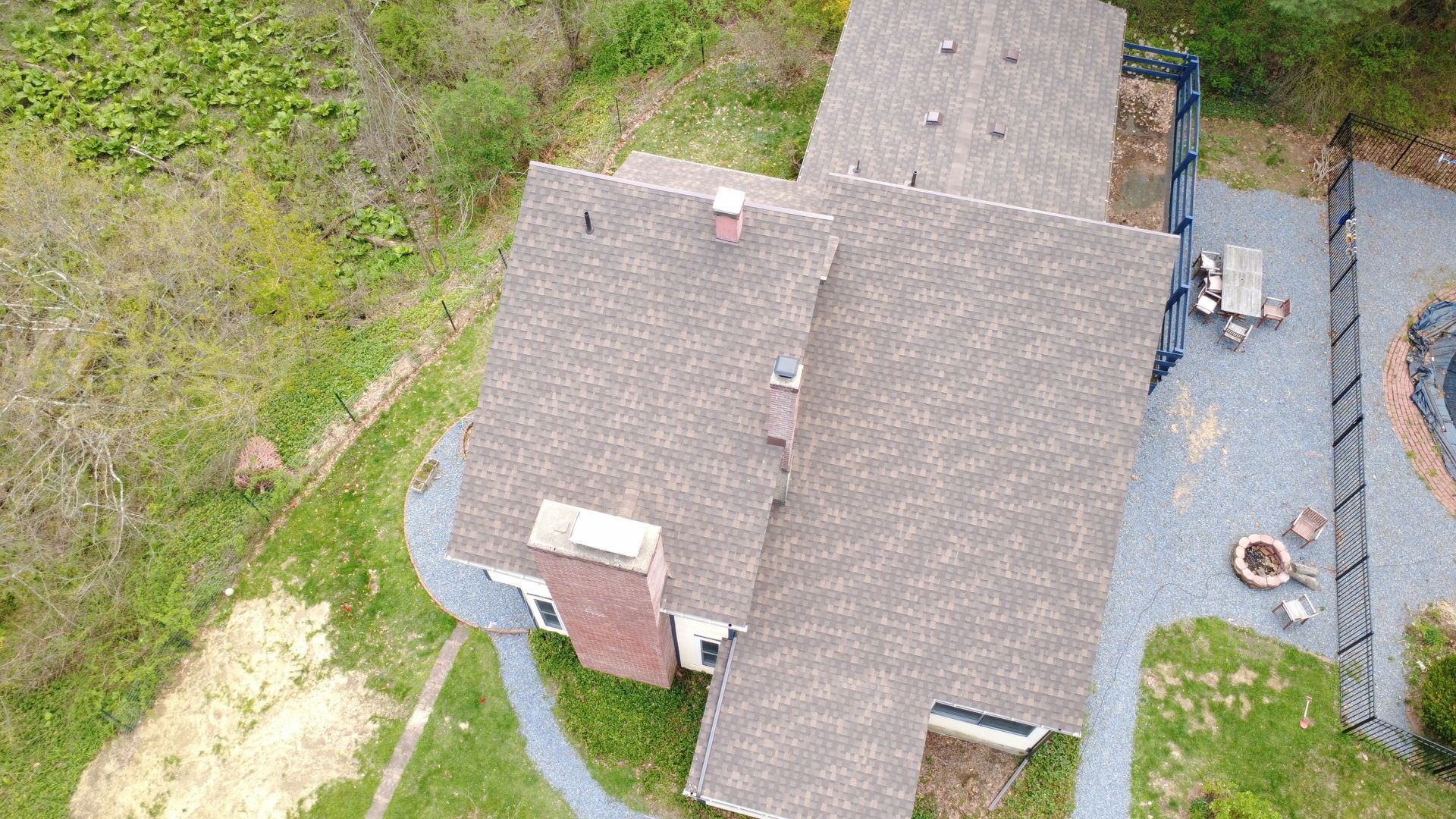 Aerial view of a house with gray shingles, a chimney, and a driveway beside grassy yard.