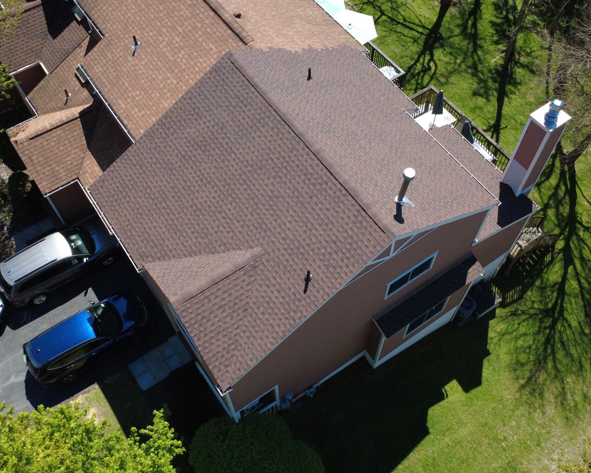 Aerial view of a brown-roofed suburban house with driveway, cars, and green yard.