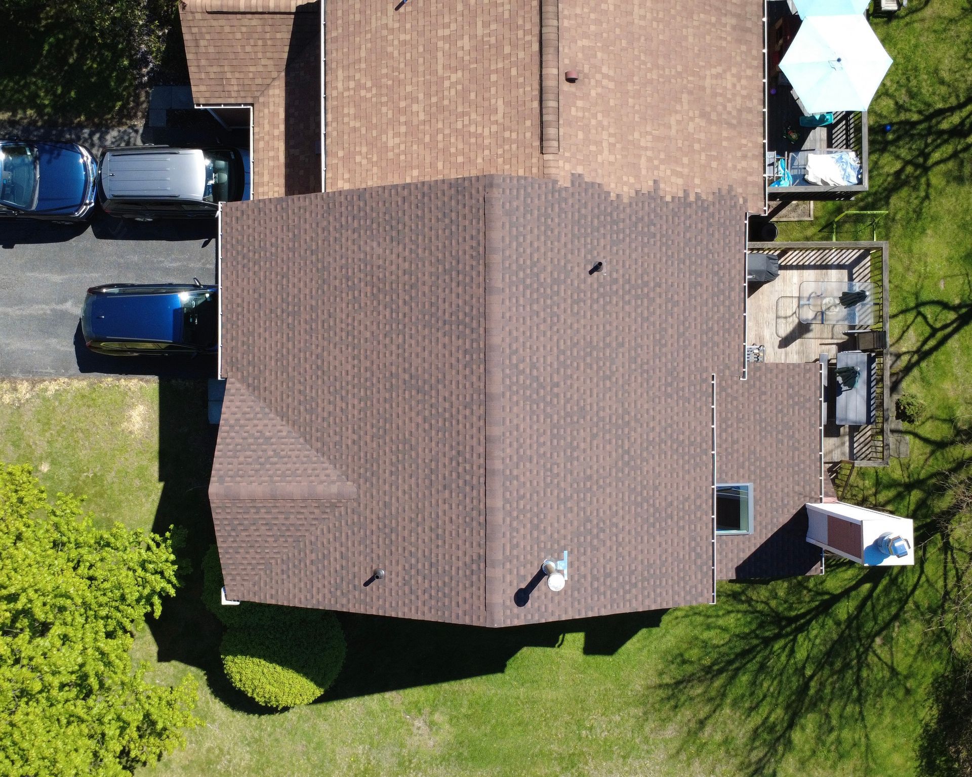 Aerial view of a house with a brown roof, driveway cars, and a backyard patio.