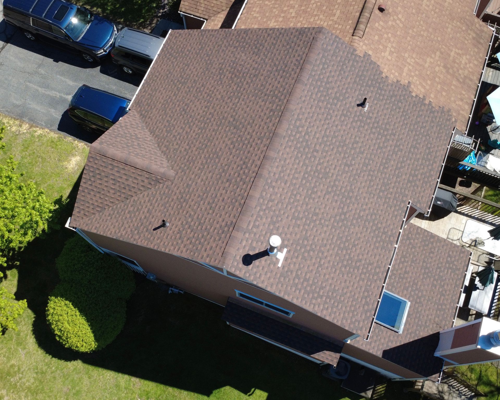 Aerial view of a brown-roofed house with a small side extension, lawn, and parked vehicles nearby.