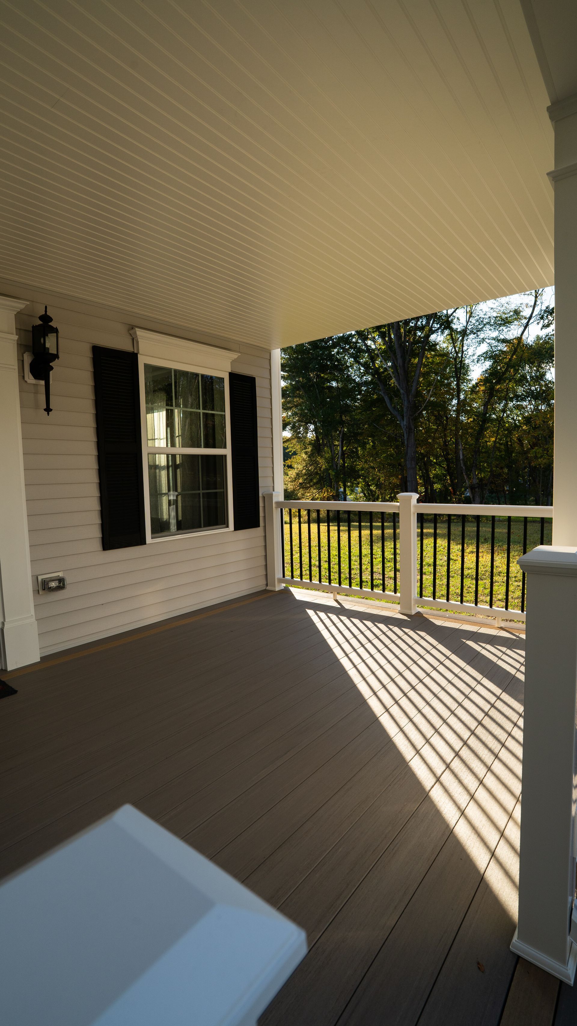 Covered front porch with beige floor, white columns, black shutters, and sunlight casting striped shadows.