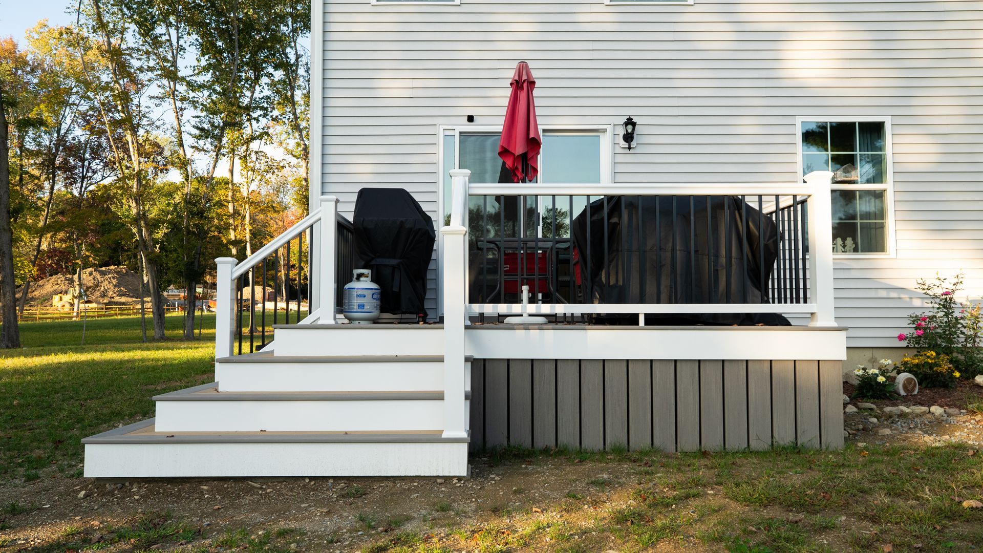 White house deck with black railing, red umbrella, grill, and white steps in a sunny yard.