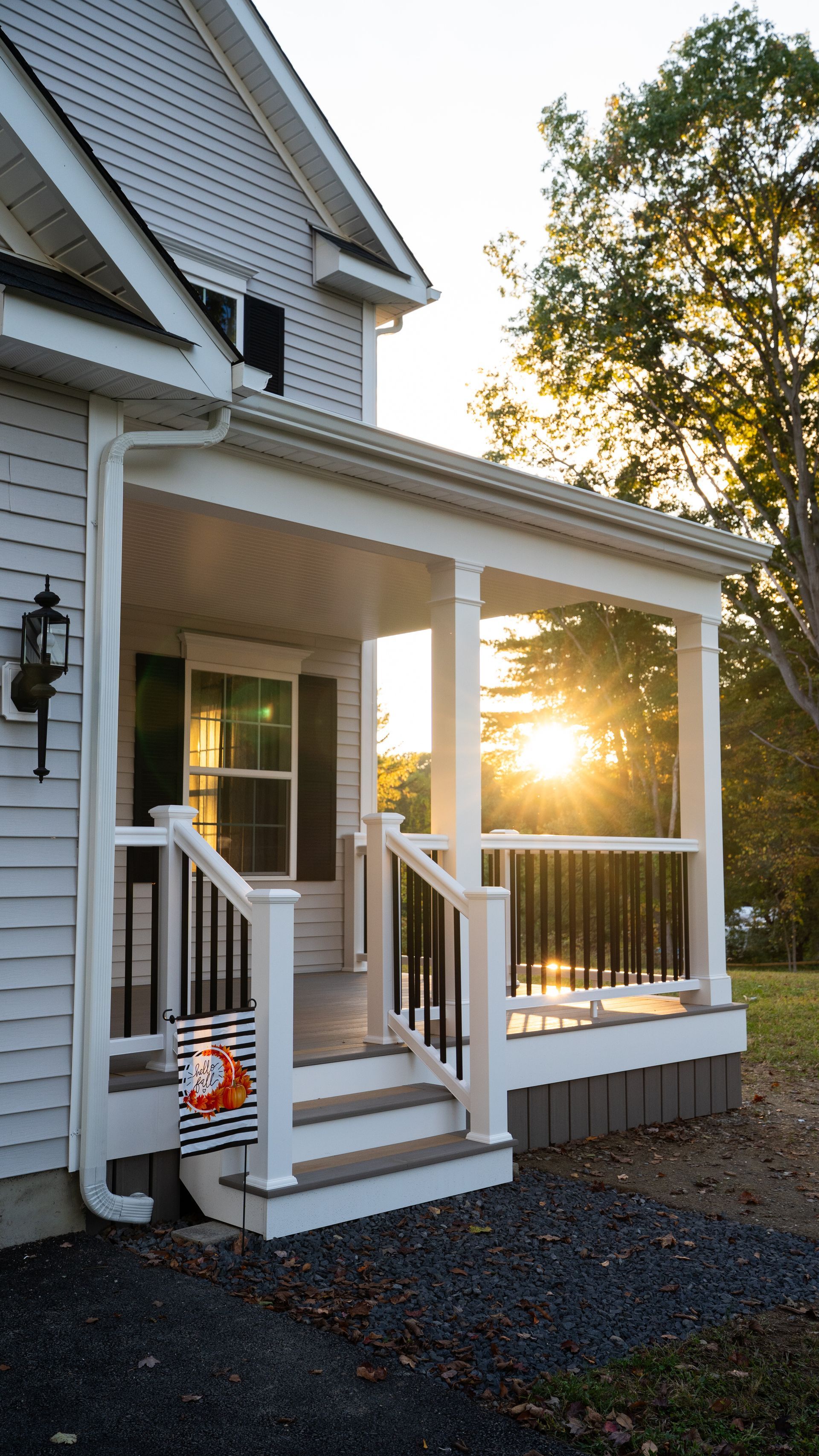 Front porch of a gray house at sunset, with white railings and warm sunlight through the trees
