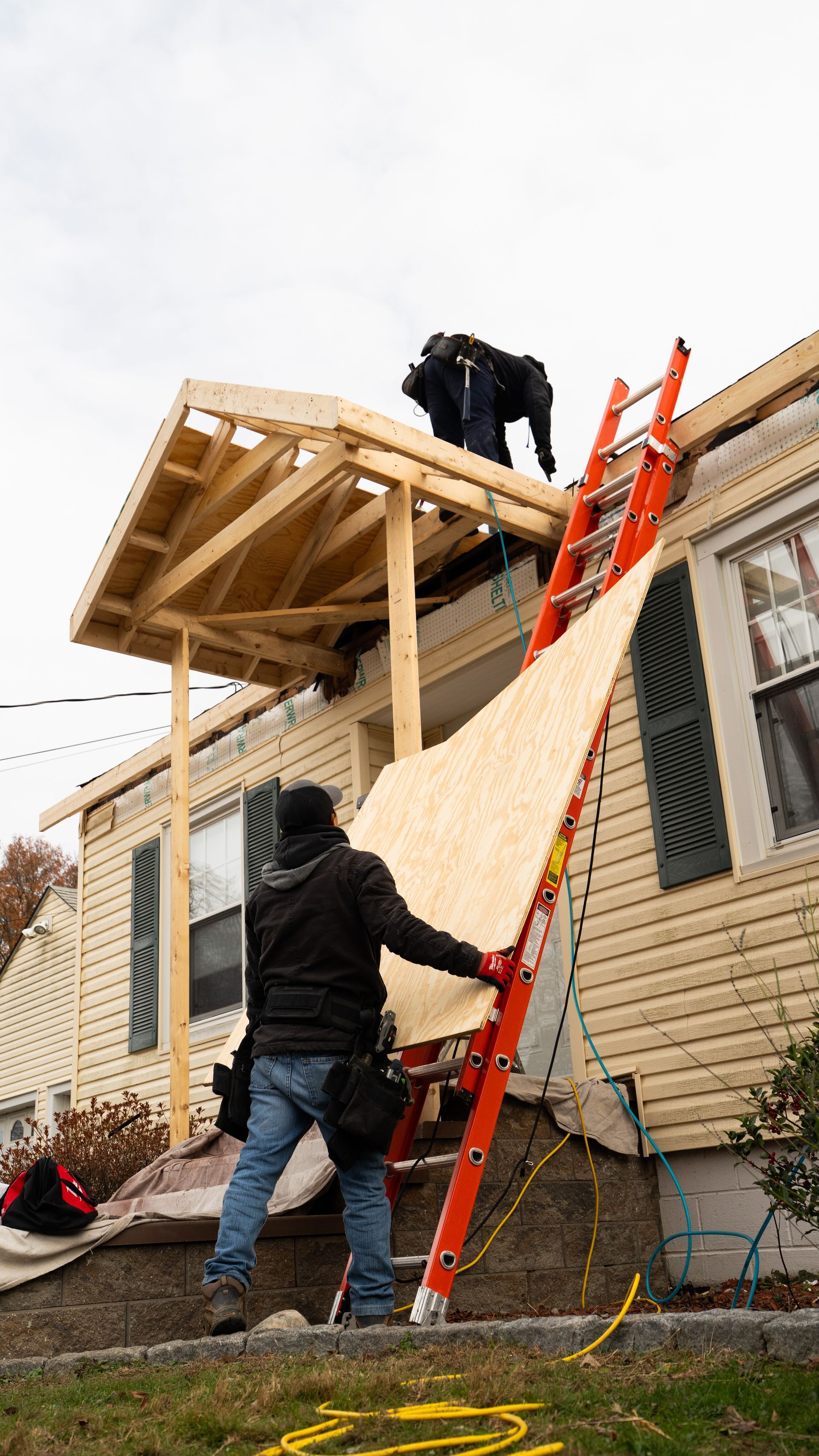Workers building a wooden roof over a brick house, with a red ladder and plywood paneling.