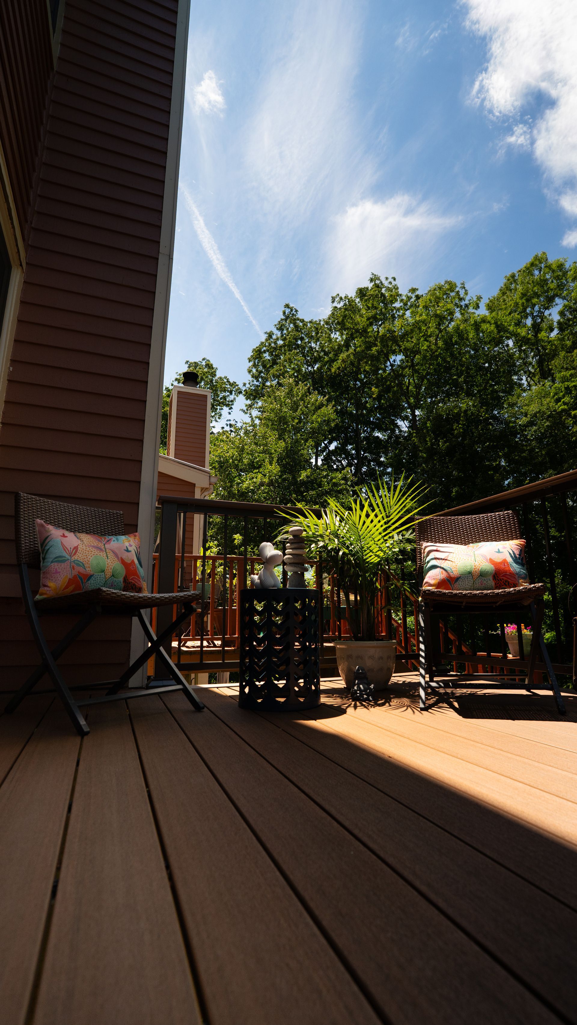 Backyard deck with two folding chairs, potted plants, and a sunny sky framed by trees and a house.